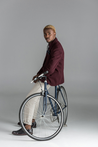 poważny człowiek afrykańskich siedząc na rowerze i patrząc na kamery na szarym tle   - Zdjęcie, obraz