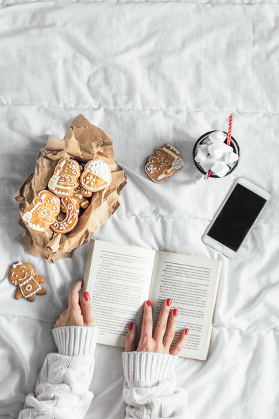 woman reading book. Top view. Smartphone, hot chocolate  with marshmallows and cookies  on white sheet - Foto, afbeelding
