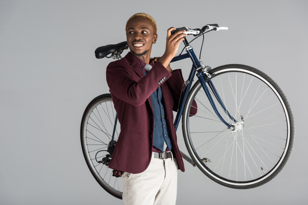 χαμογελώντας αφρικανική αμερικανική άνθρωπος που κρατά ποδήλατο στα χέρια που απομονώνονται σε γκρι   - Φωτογραφία, εικόνα