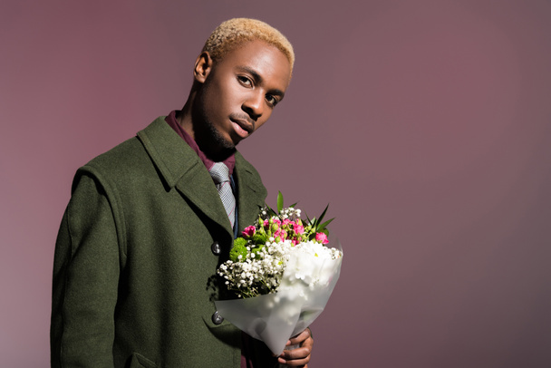 Stílusos afro-amerikai fiatalember csokor virágot látszó-on fényképezőgép, elszigetelt lila háttér - Fotó, kép