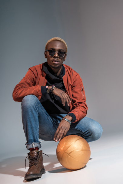 Stylish young african american man sitting with basketball ball on floor - 写真・画像