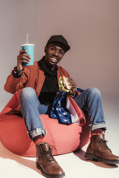 boldog fiatal afroamerikai férfi hamburger eszik, és iszik papírpohár ülve, a bab zsák széket a stúdió - Fotó, kép