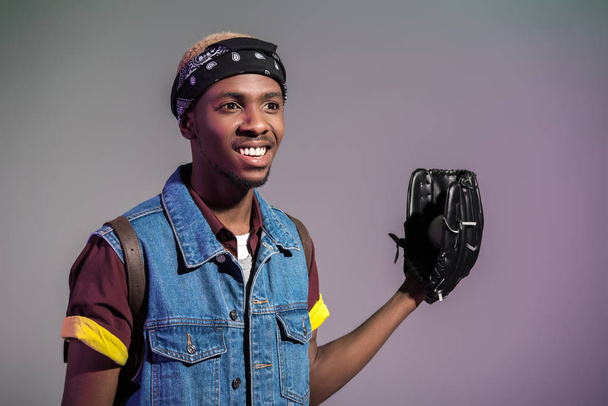 Lächelnder junger afrikanisch-amerikanischer Mann mit Baseballhandschuh, der isoliert auf grau blickt - Foto, Bild
