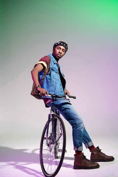 stylish young african american man sitting on bicycle and looking away on grey - Foto, Imagem