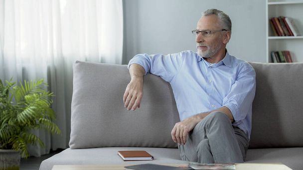 Healthy senior man wearing eyeglasses, sitting on sofa, dreaming about holiday - Foto, afbeelding