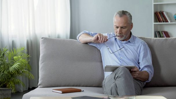 Adult male sitting on sofa and reading news on tablet, modern technologies - Foto, afbeelding