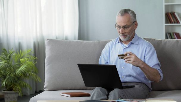 Pensionista sentado en el sofá y la inserción de número de tarjeta en el ordenador portátil, compras en línea
 - Foto, Imagen