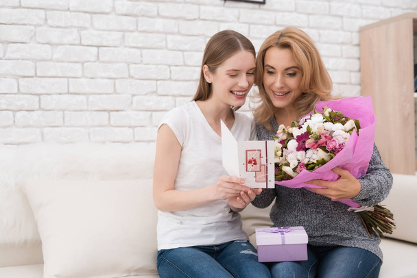 The daughter gives her mother flowers and gifts. She makes her a surprise. Mom is very surprised. They are together at home. - Photo, Image