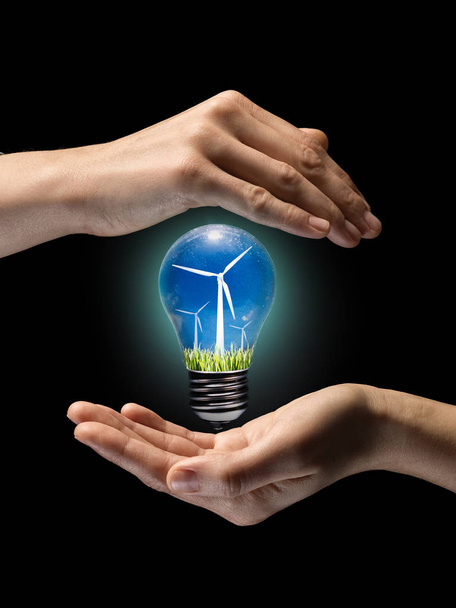 Konzept für erneuerbare Energien. Glühbirne mit Windmühle im Inneren, zwischen zwei Händen auf schwarzem Hintergrund. - Foto, Bild