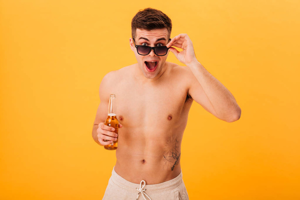 Surprised naked man in shorts and sunglasses holding beer - Фото, изображение