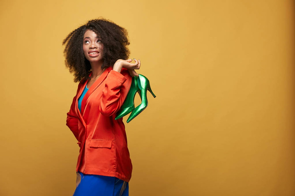 Colorful portrait of young african girl with afro hairstyle. Emotional girl wearing  orange jacket and violet latex skirt holds green heels on her fingers and posing on yellow background. Studio shot. - Фото, зображення