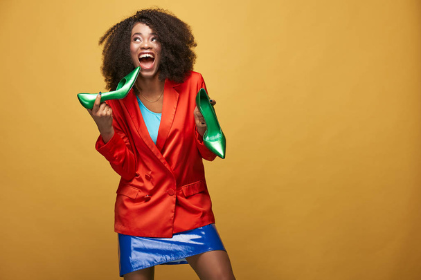 Funny colorful portrait of young african girl with afro hairstyle. Emotional girl wearing  orange jacket and violet latex skirt holds green heels in her hands screaming and posing on yellow background. Studio shot. - Fotoğraf, Görsel