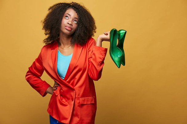 Colorful portrait of young and beautiful african girl with afro hair. Smiling girl wearing orange jacket holds green shoes on her fingers. Studio shot - Photo, Image