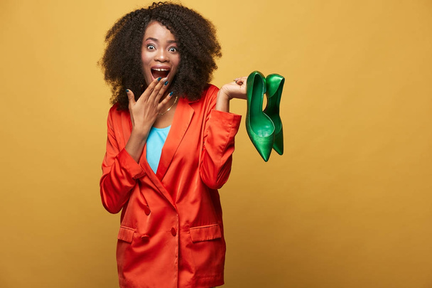 Colorful portrait of young and beautiful african girl with afro hair. Surprised girl wearing orange jacket holds green shoes on her fingers. Studio shot - Photo, Image