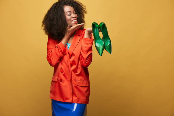 Bright and colorful portrait of young and beautiful african girl with afro hair. Smiling girl wearing orange jacket holds green shoes on her fingers. Studio shot - Photo, image
