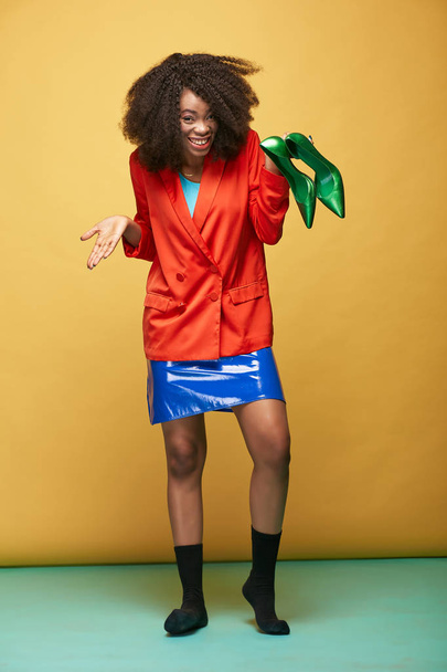 Colorful portrait of young and beautiful african girl with afro hair. Smiling girl wearing silky orange jacket, violet latex skirt and black socks  holds green shoes in her hand . Studio shot - Foto, Bild