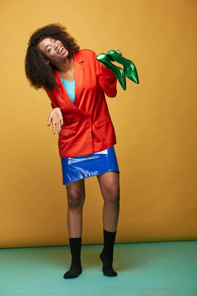 Colorido retrato de joven y hermosa chica africana con pelo afro. Chica sonriente con chaqueta naranja sedosa, falda de látex violeta y calcetines negros sostiene zapatos verdes en su mano. Captura de estudio
 - Foto, Imagen