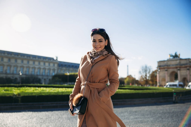 A young girl in a brown coat and scarf stands on the background of the Louvre. Autumn is sunny weather, tourists. - Photo, Image