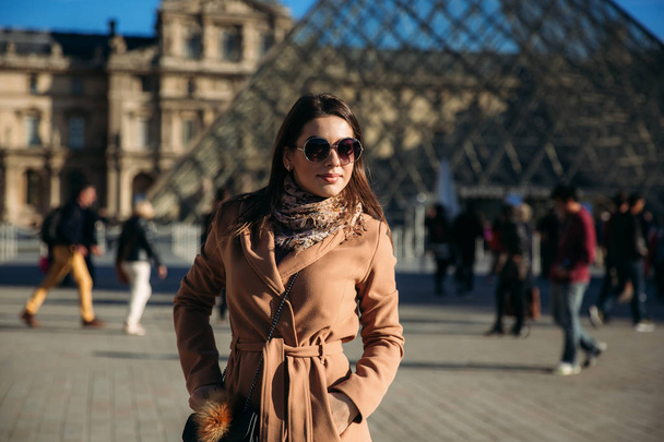 Bir genç kız bir kahverengi ceket ve eşarp Louvre arka plan üzerinde duruyor. Güneşli havalarda, turist Sonbahar geldi. - Fotoğraf, Görsel