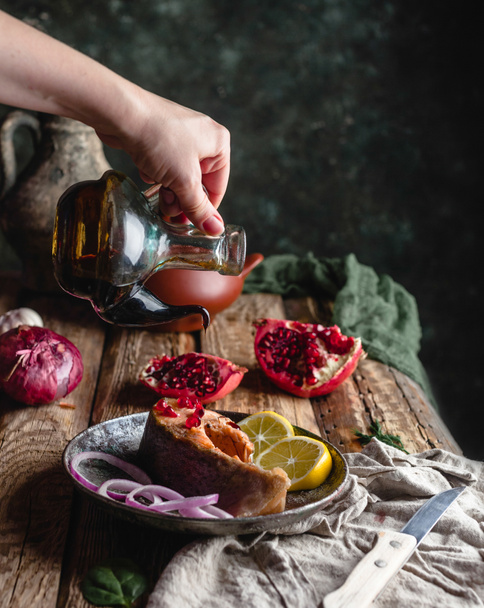 Γυναικείο χέρι ρίχνει λάδι πάνω από το ψημένο φιλέτο σολομού με το λεμόνι και το κρεμμύδι στο πιάτο με ρόδι στο ξύλινο τραπέζι - Φωτογραφία, εικόνα