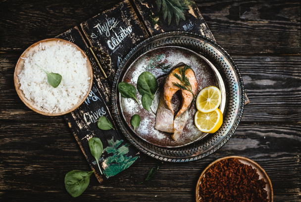 Top view of baked salmon steak with lemon on rustic metal tray with rice side dish on dark wooden table - Zdjęcie, obraz