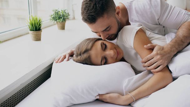 Young beautiful and loving couple wake up at the morning. Attractive man kiss and hug his wife in bed - 写真・画像