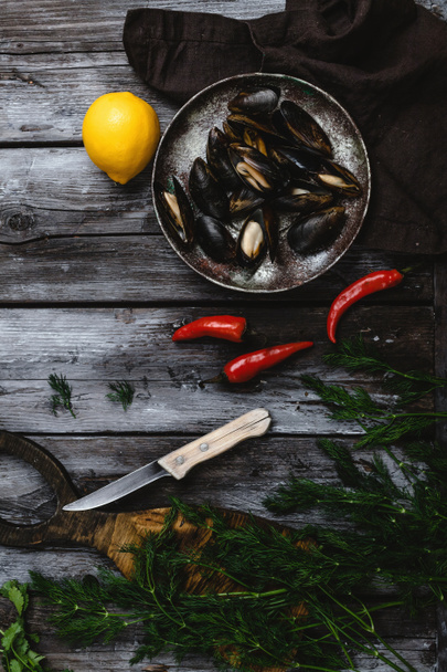 top view of delicious mussels with shells on vintage plate with herbs, chili peppers and lemon on rustic wooden table - Foto, afbeelding