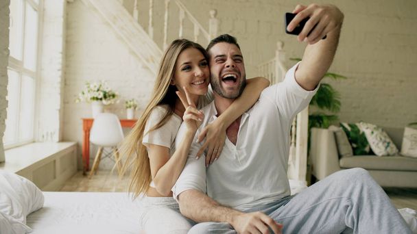 Young beautiful and loving couple take selfie picture on smartphone camera while sitting in bed at the morning - 写真・画像