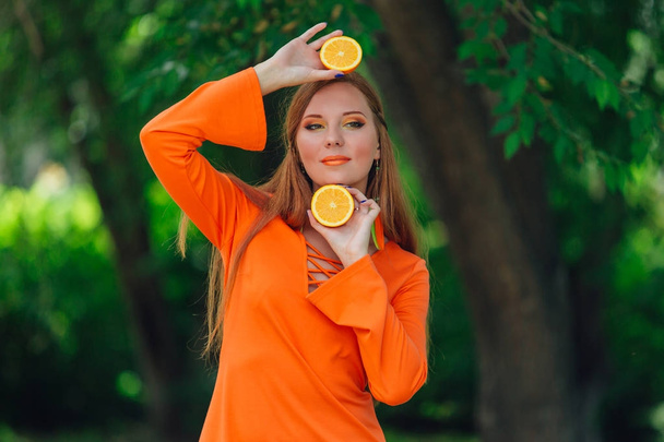 Portrait of pretty red hair woman holding two halves of juicy delicious oranges at summer green park. - Φωτογραφία, εικόνα
