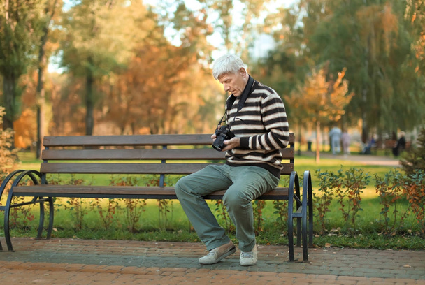 Stylish mature man with camera sitting on bench in autumn park - Photo, image