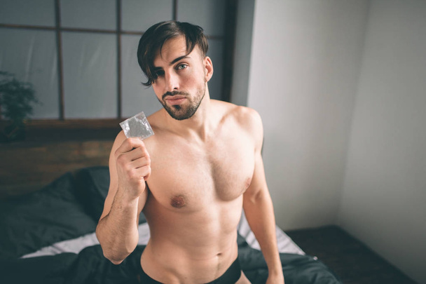 Safety sex concept. Cropped image of a naked bearded dark-hair handsome smiling man holding condom in hand on bed - Фото, изображение