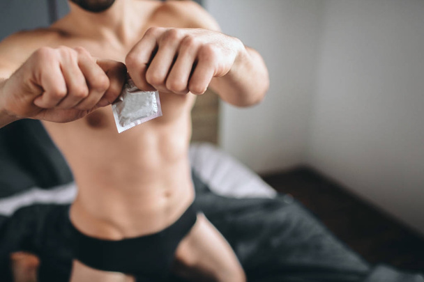 Safety sex concept. Cropped image of a naked bearded dark-hair handsome smiling man holding condom in hand on bed - Fotó, kép