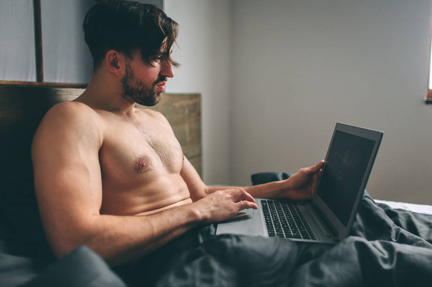 Naked bearded dark-hair Serious casual young man using laptop in bed at home - Foto, Imagem