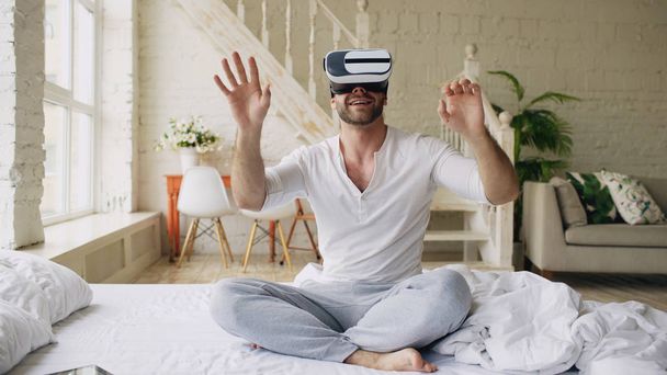 Young cheerful man wearing virtual reality headset watching 360 VR video movie sitting in the bed at home - Fotoğraf, Görsel