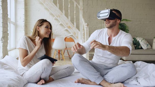 Young cute couple with tablet computer and virtual reality headset playing 360 VR video game while sitting in bed at home - Photo, Image