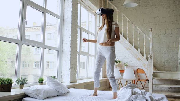 Happy smiling young woman dancing while getting experience using 360 VR headset glasses of virtual reality on bed at home - Photo, Image