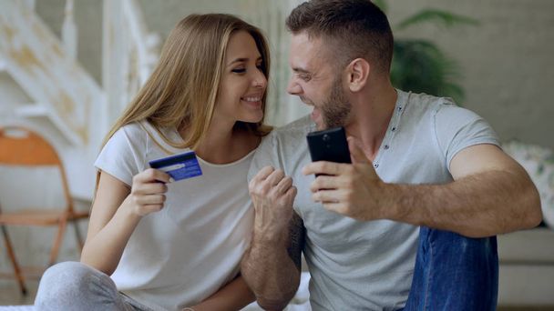 Attractive young couple with smartphone and credit card shopping on the internet sit on bed at home - Zdjęcie, obraz
