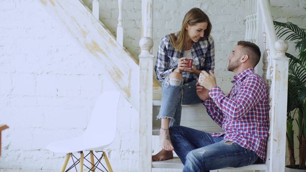 Young happy and loving couple drink tea and talking while sitting on stairs in living room at home - Foto, Imagem