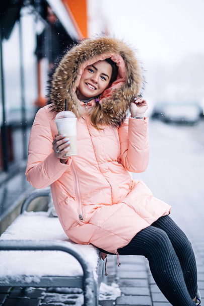 Χαμογελώντας ευτυχισμένη όμορφη γυναίκα στο ζεστό χειμώνα σακάκι υπαίθρια απολαμβάνει ζεστό καφέ, λοιπόν φόρεσαν γάντια και κουκούλα, χιονίζει καιρός - Φωτογραφία, εικόνα