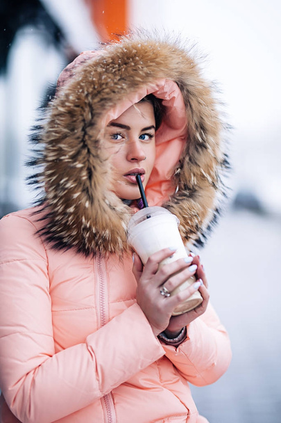 Χαμογελώντας ευτυχισμένη όμορφη γυναίκα στο ζεστό χειμώνα σακάκι υπαίθρια απολαμβάνει ζεστό καφέ, λοιπόν φόρεσαν γάντια και κουκούλα, χιονίζει καιρός - Φωτογραφία, εικόνα