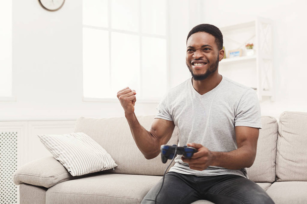 Χαρούμενος νεαρός άνδρας Αφροαμερικανός στο σπίτι παίζοντας βιντεοπαιχνίδια και νίκες - Φωτογραφία, εικόνα