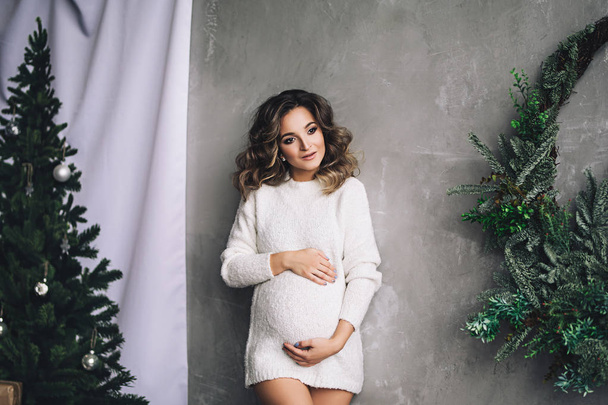 Caucasian pregnant woman with make up and curly hair in warm white sweater holds her belly, portrait of future mother, happy pregnancy, fashion portrait, stylish pregnancy - Foto, afbeelding