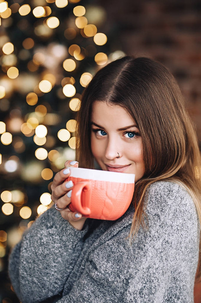 Feliz mujer sonriente en el suéter bebe chocolate caliente cerca del brillante árbol de Navidad en su sala de estar, víspera de Navidad, comodidad en el hogar y el estado de ánimo de Año Nuevo, bebida caliente, mujer bastante atractiva
 - Foto, imagen