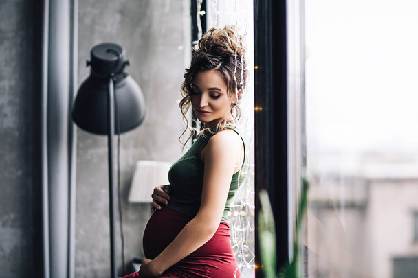 kaukasische schwangere Frau mit Make-up und lockigem Haar sitzt neben den großen Fenstern in stilvollen skandinavischen Interieurs, Porträt der zukünftigen Mutter, glückliche Schwangerschaft, Modeporträt, stilvolle Schwangerschaft - Foto, Bild