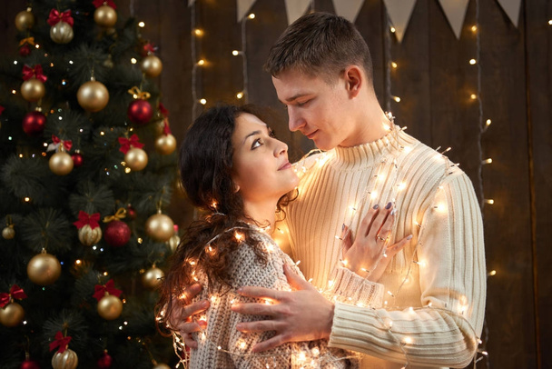 young couple together in christmas lights and decoration, dressed in white, fir tree on dark wooden background, romantic evening, winter holiday concept - Zdjęcie, obraz