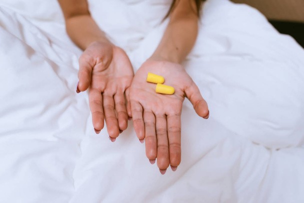 the girl sits in bed under a white blanket, in hands yellow earplugs against street noise - Photo, Image