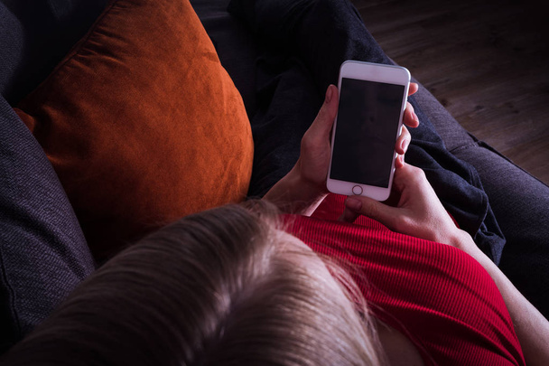 A woman lies on the couch in the evening and holds a mobile phone - Fotoğraf, Görsel