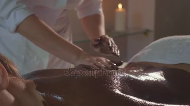 Beautician doing massage with chocolate to young beautiful woman at spa salon - Felvétel, videó