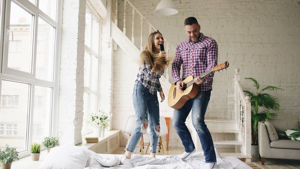 Funny happy and loving couple dance on bed singing with tv controller and playing guitar. Man and woman have fun during their holiday at home - 写真・画像