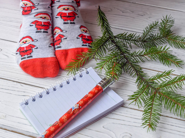 A festive Christmas photo with feminine feet in bright red socks, a fir twig, a note book and a pencil on a white wooden floor  - Fotó, kép